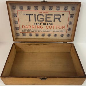 May include: An open, rectangular wooden box with a hinged lid. The inside of the lid features a printed label with the text "TIGER" and "FAST BLACK DARNING COTTON." The box is empty, showing its natural wood grain.