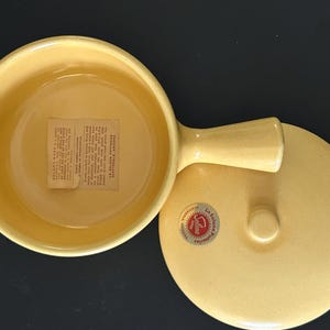 May include: A yellow ceramic casserole dish with a handle and lid. The dish has a round shape and a smooth surface. A sticker is visible on the lid, and a label is inside the dish. The dish is likely vintage.