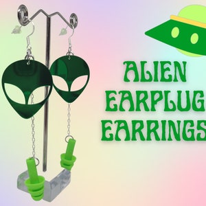 May include: Green alien head earrings with a dangling green earplug charm. The earrings are on a display stand with the text "ALIEN EARPLUG EARRINGS" in green letters.