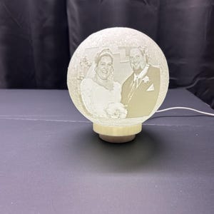 May include: A white globe lamp with a 3D engraved image of a bride and groom. The lamp has a white base and a white cord.