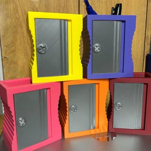May include: A collection of colorful, 3D-printed desk organizers. Each organizer features a rectangular design with a gray door and a decorative knob. The organizers are in various colors, including yellow, purple, pink, orange, and red. Some contain pens.