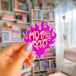 May include: A pink sticker in the shape of a heart with flames around it. The sticker has the words "HOT WITH THE PLOTS" in a stylized font. The sticker is held in front of a blurred background of bookshelves.