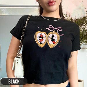 Op de afbeelding: Zwart getailleerd T-shirt met een afbeelding van Mickey Mouse en Minnie Mouse die elkaar kussen in een hartvormig medaillon. Het medaillon is versierd met een roze lint.