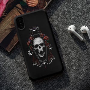 May include: Black phone case featuring a skull design within a coffin, adorned with red roses and bats. The case is next to a pair of white wireless earbuds. The design is a gothic or Halloween theme.