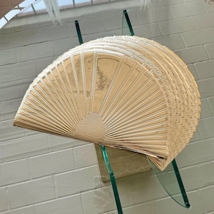 May include: A gold-colored, fan-shaped decorative object, possibly a placemat or serving piece, is displayed on a glass table. The object has a textured surface with radial lines emanating from a central point. The table has a clear glass top and green glass supports.