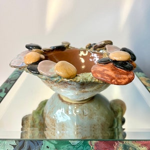 Könnte beinhalten: Eine Keramikschale mit hellblauer Glasur und braunem Innenraum. Die Schale ist mit kleinen, runden, braunen, hellbraunen und schwarzen Steinen verziert. Die Schale steht auf einer Spiegelfläche mit einem Blumendesign.