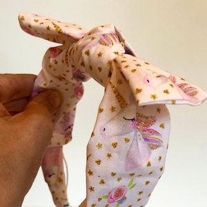 May include: A pink fabric headband with a bow, decorated with gold stars and unicorn illustrations. The headband is connected to two white hearing aid devices. The fabric features pink roses and gold glitter accents.