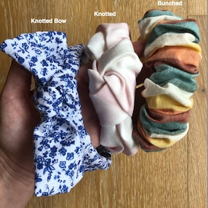 以下が含まれることがあります： 異なるスタイルの3つの布製のヘアバンド。青と白の花柄の結び目のあるボウヘアバンド、薄いピンク色の結び目のあるヘアバンド、緑、黄色、茶色のストライプ柄の束ねられたヘアバンド。