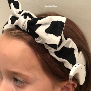 Può includere: Un cerchietto nero e bianco con stampa a mucca con un fiocco annodato. Il cerchietto è indossato da una persona con i capelli castani e un apparecchio acustico.