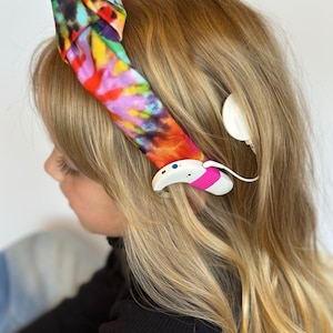 May include: A young person with long blonde hair wearing a colorful headband and a hearing aid.