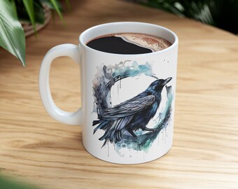 May include: White ceramic mug with a black raven illustration in a watercolor circle design. The raven is perched on a branch with its wings spread. The mug is filled with coffee.