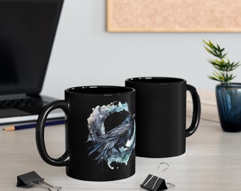 May include: Two black ceramic coffee mugs. One mug features a watercolor illustration of a raven perched within a circular design with blue and gray accents. The other mug is plain black. Both mugs have a black handle.