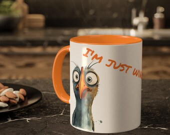 May include: White ceramic mug with an orange interior and handle. The mug features a cartoon bird illustration with the text "I'M JUST WINGING IT" in orange. The mug is on a black surface.