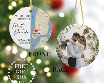 May include: A round, white ceramic ornament with a map of New York City on the front. The map has a red heart marking the location of "New York". The text "WHERE SHE SAID 'YES'" is written above the map. Below the map, the text "Matt & Pamela ENGAGED 06.22.2024" is written. The back of the ornament has a photo of a couple embracing in a floral setting. The text "FREE GIFT BOX" is written below the ornament.