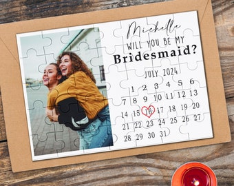 May include: A puzzle card with a photo of two women smiling. The card has the text "Michelle, Will you be my Bridesmaid?" and a calendar with July 2024 highlighted with a red heart on the 16th.