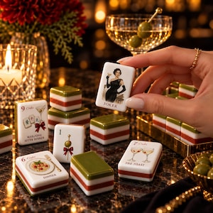 May include: A set of white, red, and green mahjong tiles featuring illustrations of cocktails, food, and a woman in a black dress. One tile is held up, displaying the word JOKER. Other tiles show text including 9 Craks, MAHJONG AFTER DINNER, 6 Craks, DINNER, 1, 7 Craks, and THE TOAST.