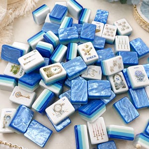 May include: A collection of mahjong tiles featuring a mix of solid blue and green striped designs and white tiles with intricate illustrations, including a Joker tile, floral patterns, keys, clocks, and teacups, all arranged in a scattered pile.