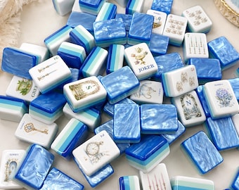 May include: A collection of mahjong tiles featuring a mix of solid blue and green striped designs and white tiles with intricate illustrations, including a Joker tile, floral patterns, keys, clocks, and teacups, all arranged in a scattered pile.