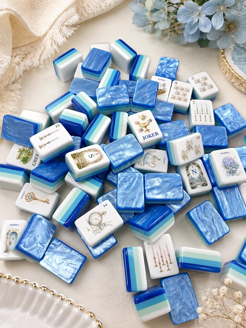 K&ouml;nnte beinhalten: Eine Sammlung von Mahjong-Steinen mit einer Mischung aus blau-gr&uuml;n gestreiften Designs und wei&szlig;en Steinen mit detaillierten Illustrationen, darunter ein Joker-Stein, Blumenmuster, Schl&uuml;ssel, Uhren und Teetassen, die in einem Haufen liegen.