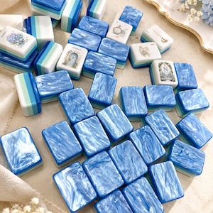 K&ouml;nnte beinhalten: Eine Sammlung rechteckiger Mahjong-Spielsteine mit einer marmorierten blau-wei&szlig;en Oberfl&auml;che. Einige Steine zeigen Illustrationen von Blumen, einer Uhr, einer Teetasse, einer T&uuml;r und dem Buchstaben N, andere ein blau-wei&szlig; gestreiftes Muster.