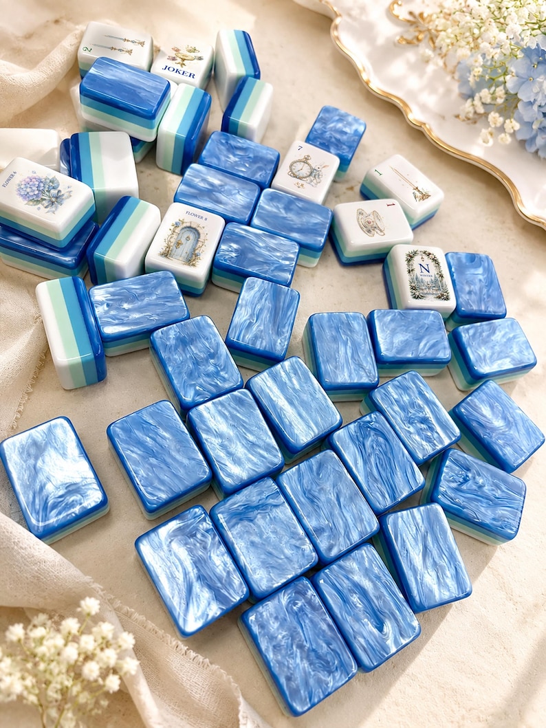 K&ouml;nnte beinhalten: Eine Sammlung rechteckiger Mahjong-Spielsteine mit einer marmorierten blau-wei&szlig;en Oberfl&auml;che. Einige Steine zeigen Illustrationen von Blumen, einer Uhr, einer Teetasse, einer T&uuml;r und dem Buchstaben N, andere ein blau-wei&szlig; gestreiftes Muster.