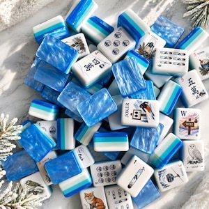 Peut inclure: Un ensemble de tuiles de mah-jong avec des dos marbrés bleu et blanc et des faces blanches illustrées de motifs variés comme un renard, un hibou, un skieur, une lanterne, un téléphérique, des caractères chinois et des chiffres.