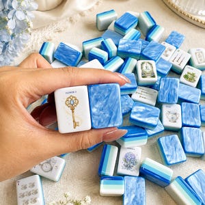 K&ouml;nnte beinhalten: Eine Sammlung rechteckiger Mahjong-Spielsteine mit blau-wei&szlig;en Marmormustern, einfarbigen blau-wei&szlig;en Streifen und bedruckten Motiven, darunter ein goldener Schl&uuml;ssel mit der Aufschrift FLOWER 3, ein Blumenstrau&szlig; und Teetassen.
