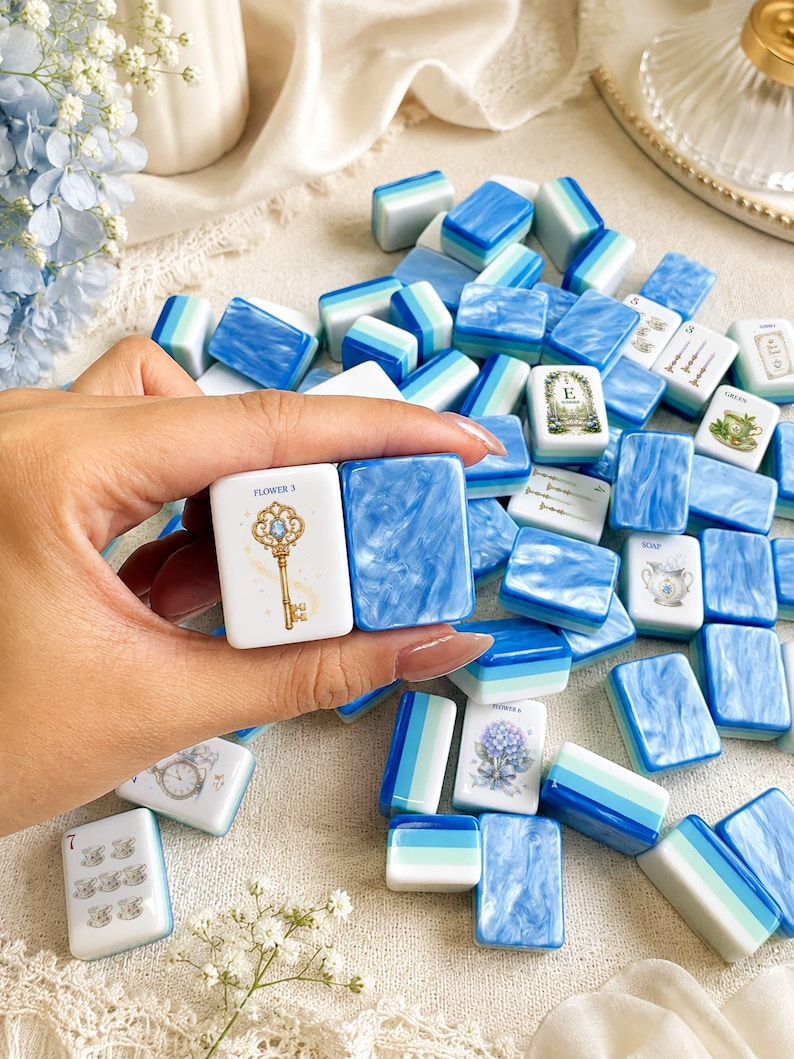 K&ouml;nnte beinhalten: Eine Sammlung rechteckiger Mahjong-Spielsteine mit blau-wei&szlig;en Marmormustern, einfarbigen blau-wei&szlig;en Streifen und bedruckten Motiven, darunter ein goldener Schl&uuml;ssel mit der Aufschrift FLOWER 3, ein Blumenstrau&szlig; und Teetassen.
