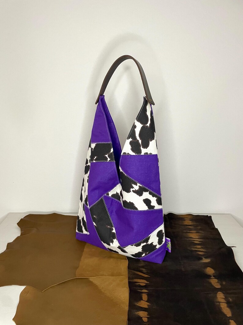 Op de afbeelding: Een paarse en zwarte patchwork tote bag met een koe-print ontwerp. De tas heeft een bruin leren handvat.