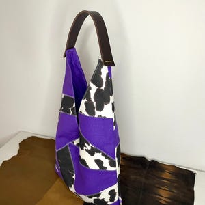 Op de afbeelding: Paarse en witte patchwork tote bag met een bruin leren hengsel. De tas heeft een koe-print patroon en is gemaakt van verschillende stoffen.