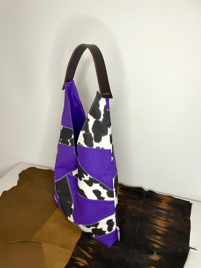 Op de afbeelding: Paarse en witte patchwork tote bag met een bruin leren hengsel. De tas heeft een koe-print patroon en is gemaakt van verschillende stoffen.