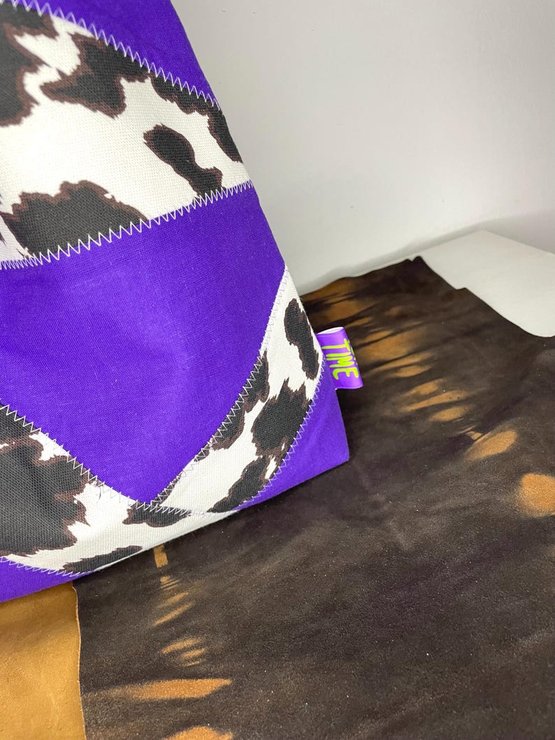 Op de afbeelding: Een paarse tote bag met een patchwork ontwerp met een wit en bruin koeienprint. De tas heeft een groen label met het woord "TIME" erop.