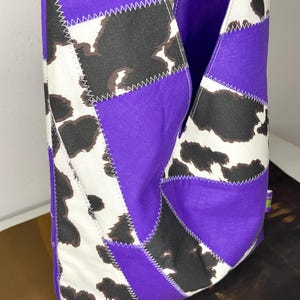 Op de afbeelding: Een paarse en witte patchwork tote bag met een koe-print ontwerp. De tas is gemaakt van stof en heeft een unieke, geometrische vorm.