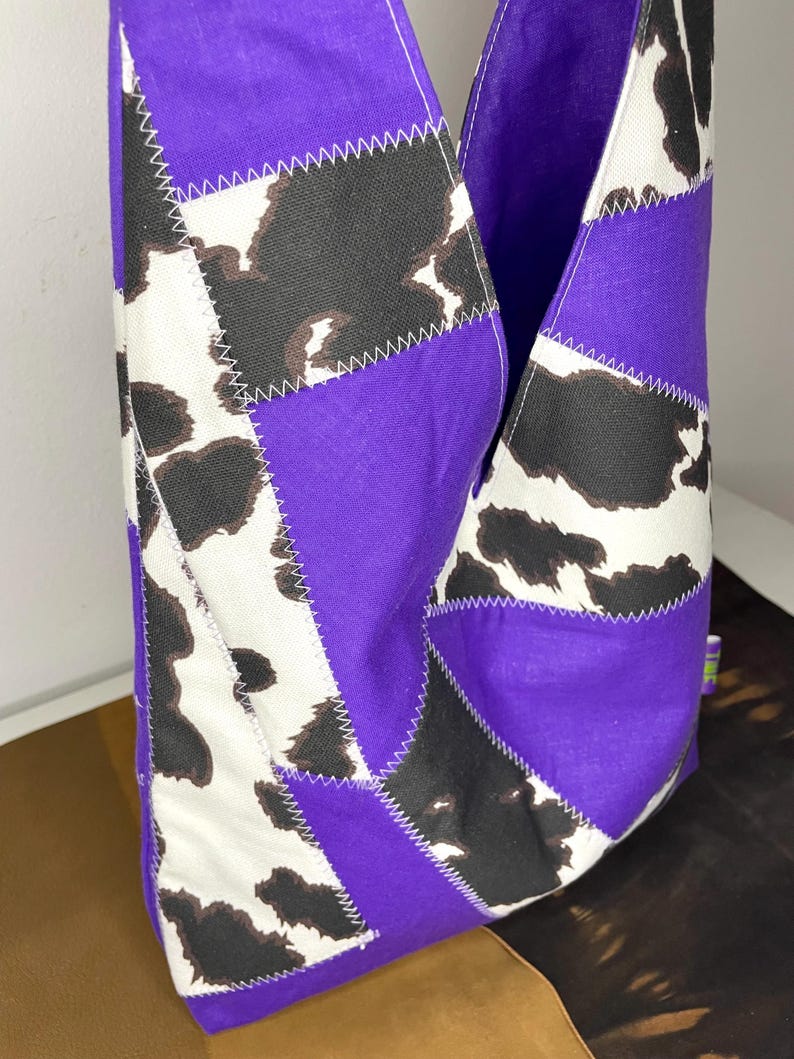 Op de afbeelding: Een paarse en witte patchwork tote bag met een koe-print ontwerp. De tas is gemaakt van stof en heeft een unieke, geometrische vorm.