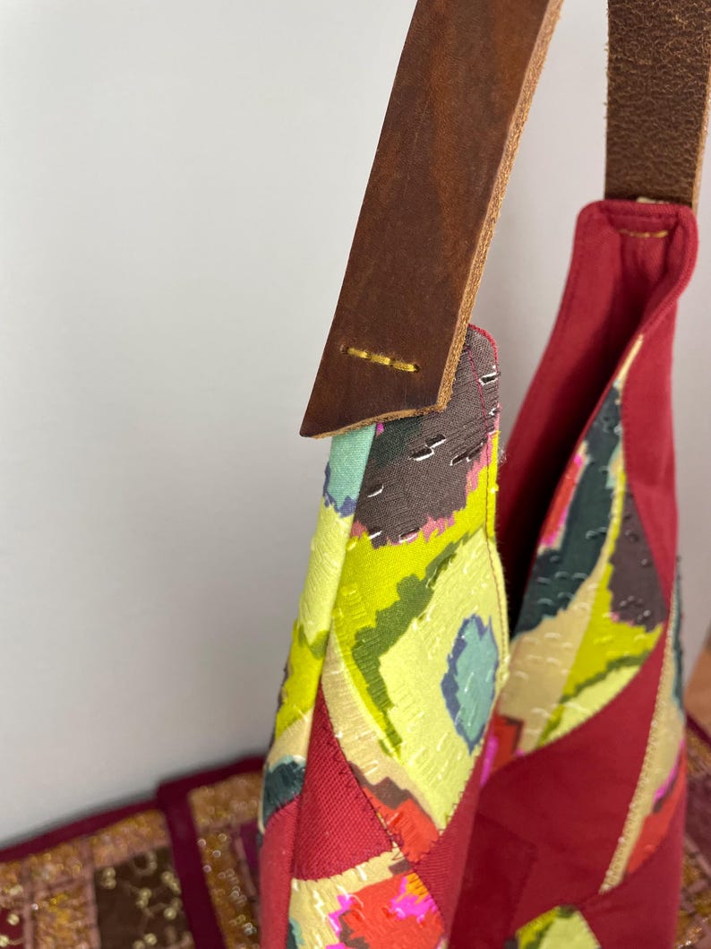 May include: A red, green and yellow patchwork tote bag with a brown leather strap. The bag has a geometric pattern and is made of different fabrics.
