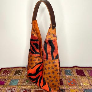 Pode incluir: Uma bolsa de ombro de couro marrom com um design de patchwork. A bolsa &eacute; feita de diferentes tecidos, incluindo estampa de zebra laranja, preto e branco, e um padr&atilde;o floral. A bolsa tem uma al&ccedil;a longa de couro marrom.