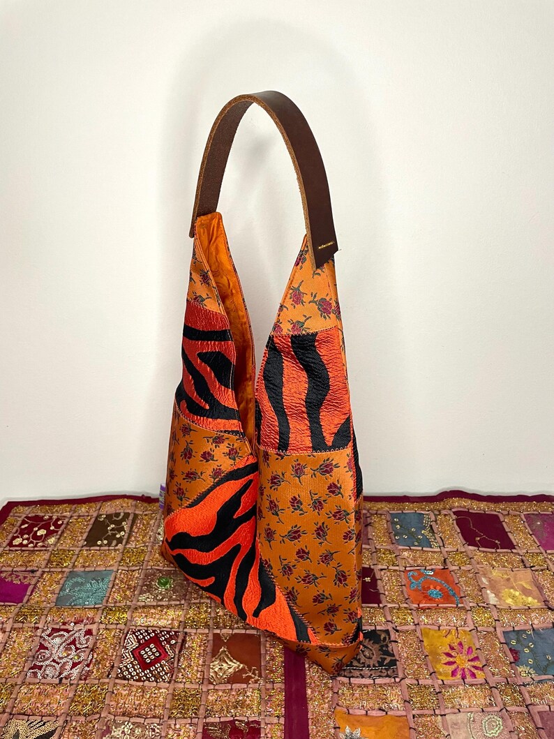 Pode incluir: Uma bolsa de ombro de couro marrom com um design de patchwork. A bolsa &eacute; feita de diferentes tecidos, incluindo estampa de zebra laranja, preto e branco, e um padr&atilde;o floral. A bolsa tem uma al&ccedil;a longa de couro marrom.