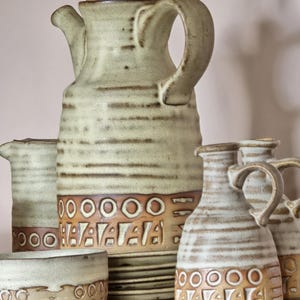 May include: A collection of handcrafted ceramic pottery in shades of green and brown. The set includes a tall teapot with a lid, a creamer, a small bowl, and two handled pitchers. The pottery features a textured surface with horizontal lines and geometric patterns.