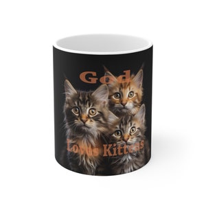 Puede incluir: Una taza de café negra con una imagen de tres gatitos. Los gatitos están mirando a la cámara. El texto "God Loves Kittens" está impreso en la taza en naranja.