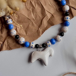 May include: A handcrafted necklace featuring a white ceramic horse pendant. The necklace has a beaded design with blue, white, and black beads. The beads are interspersed with silver-toned metal accents, creating a unique and artistic accessory.
