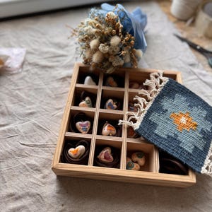 May include: A wooden box with multiple compartments, each holding a heart-shaped item. The hearts have various designs and colors. A small bouquet of dried flowers wrapped in blue paper sits above the box. A small, blue and orange patterned textile is on the right.