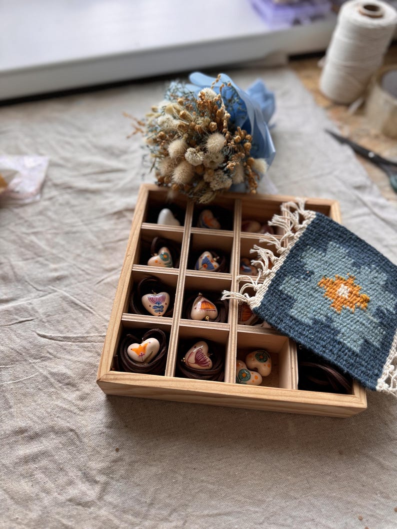 May include: A wooden box with multiple compartments, each holding a heart-shaped item. The hearts have various designs and colors. A small bouquet of dried flowers wrapped in blue paper sits above the box. A small, blue and orange patterned textile is on the right.