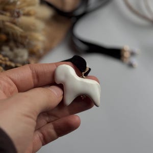 May include: A white ceramic dog pendant on a black velvet choker necklace. The dog pendant is a stylized, minimalist design. The necklace has gold-colored accents and a clasp. The background is blurred, with neutral tones.