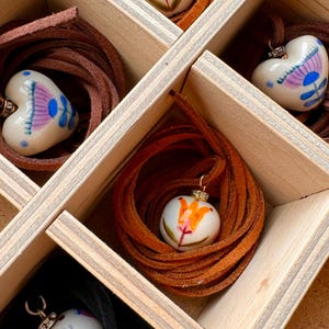 May include: A wooden box with multiple compartments, each containing a heart-shaped pendant with a painted floral design. The pendants are white with colorful illustrations and are strung on brown leather cords. The box is made of light-colored wood.
