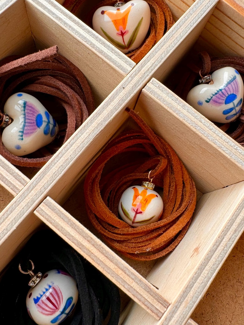 May include: A wooden box with multiple compartments, each containing a heart-shaped pendant with a painted floral design. The pendants are white with colorful illustrations and are strung on brown leather cords. The box is made of light-colored wood.