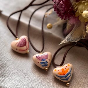 May include: Three heart-shaped pendants with hand-painted floral designs. Each pendant is strung on a brown cord. The pendants are in shades of pink, blue, orange, and white. A bouquet of dried flowers is in the background.