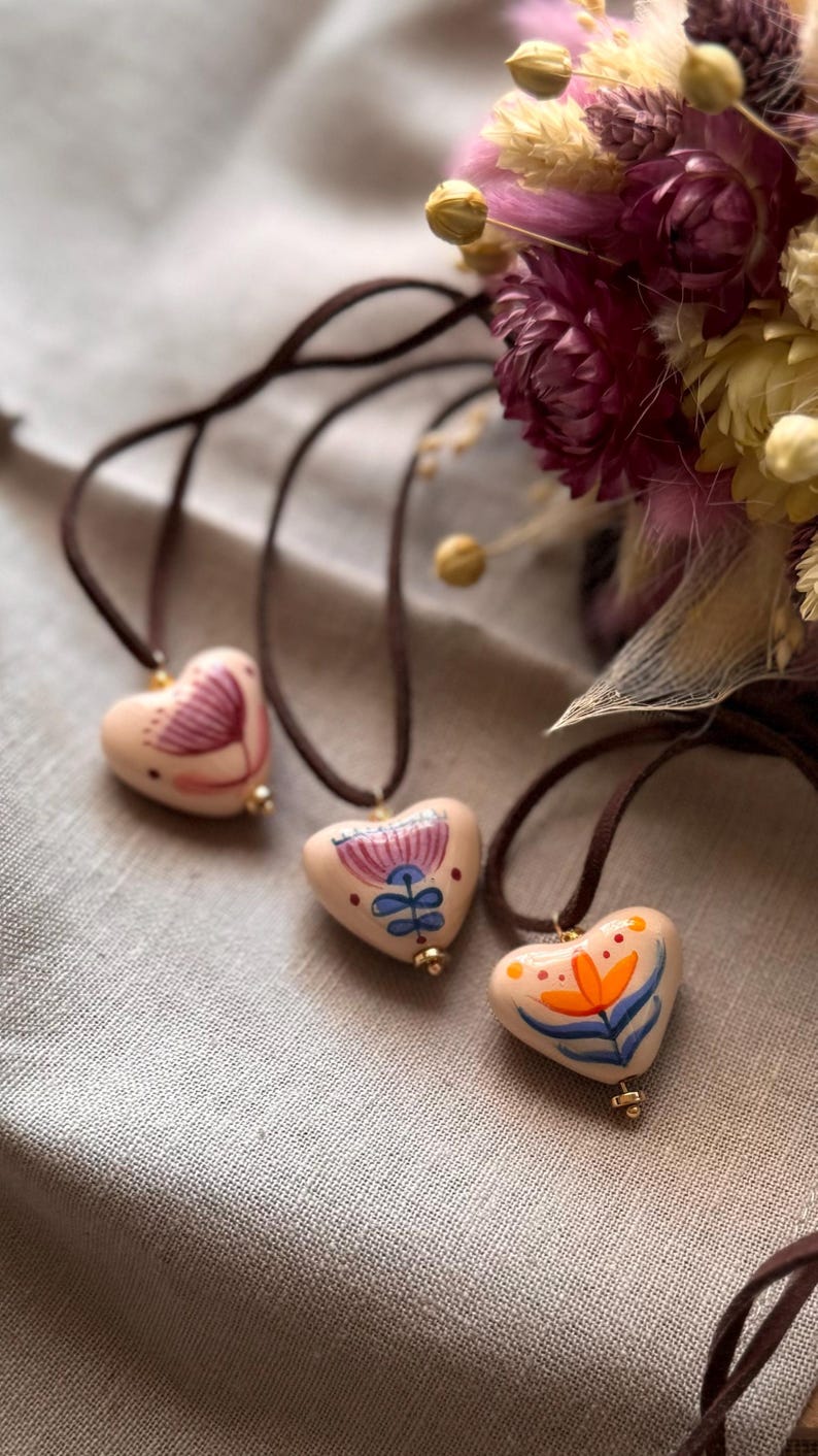 May include: Three heart-shaped pendants with hand-painted floral designs. Each pendant is strung on a brown cord. The pendants are in shades of pink, blue, orange, and white. A bouquet of dried flowers is in the background.