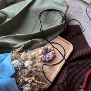 May include: A handmade necklace with heart-shaped pendants, each adorned with colorful floral designs. The necklace rests on a wooden tray, alongside a bouquet of dried flowers wrapped in blue paper. A black cord and red pliers are also visible.