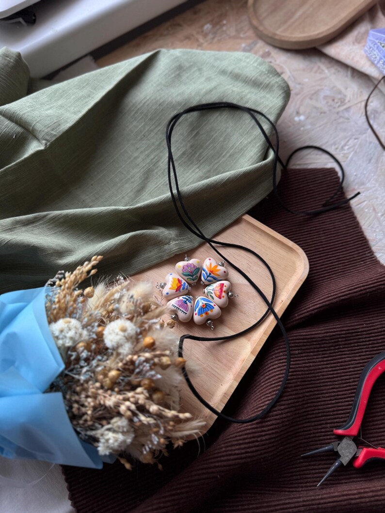 May include: A handmade necklace with heart-shaped pendants, each adorned with colorful floral designs. The necklace rests on a wooden tray, alongside a bouquet of dried flowers wrapped in blue paper. A black cord and red pliers are also visible.