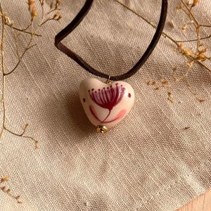 May include: A heart-shaped pendant necklace with a dark brown suede cord. The pendant is off-white with a hand-painted floral design in shades of pink and burgundy. A small gold-colored bead accents the bottom of the pendant.